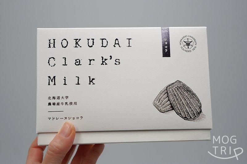 北海道大学の敷地内にある北大マルシェ の「HOKUDAI Clark's Milk マドレーヌ ショコラ」の箱を手に持っている様子