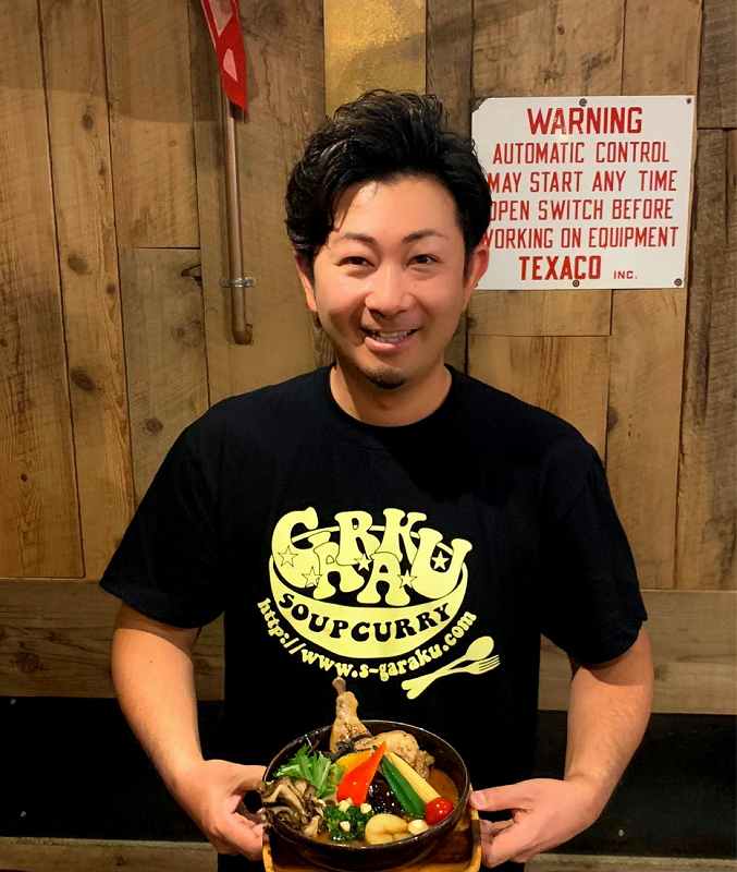 ガラクのオーナー 福生 雅寿さんがスープカレーを持って笑顔で立っている